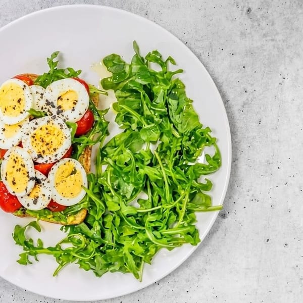 Avocado toast with sliced hard-boiled eggs, cherry tomatoes, and a side of fresh arugula.