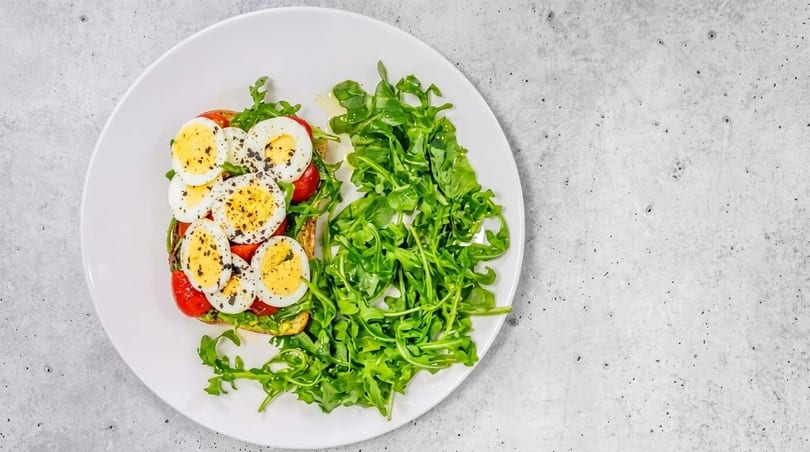 Avocado toast with sliced hard-boiled eggs, cherry tomatoes, and a side of fresh arugula.